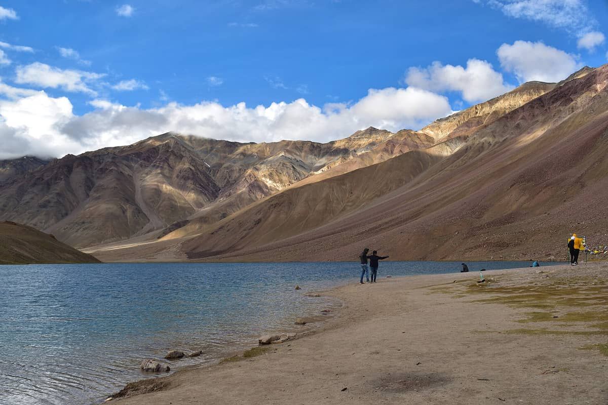 Travellers on the pebble shore of Chandratal lake below brown mountains